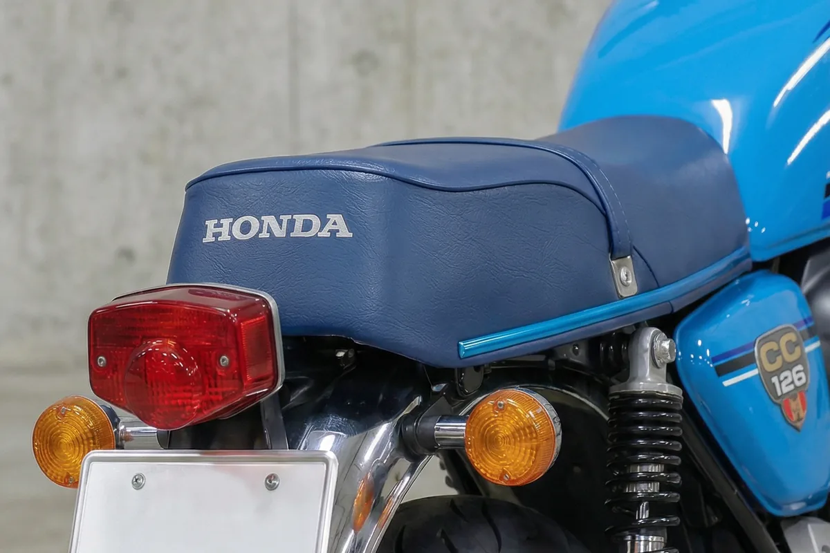 Parte traseira da motocicleta com banco azul Honda, lanterna traseira vermelha e piscas laranjas.